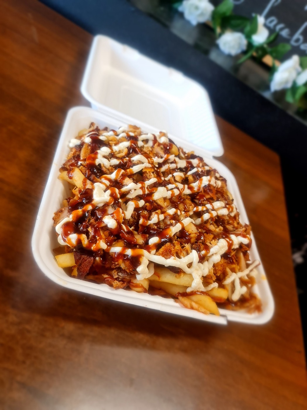 A white takeaway container of hot chips topped with shredded meat, drizzled with white and brown sauces