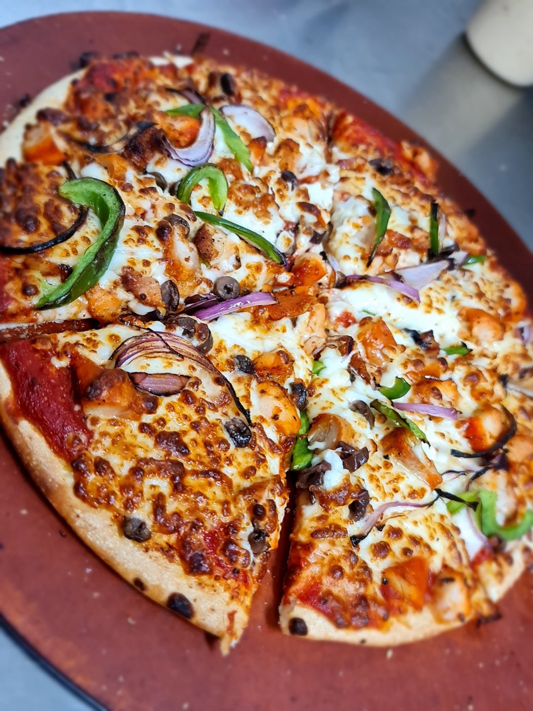 A freshly baked pizza on a round board, topped with melted cheese, chicken pieces, green capsicum, red onion and olives, with two slices cut