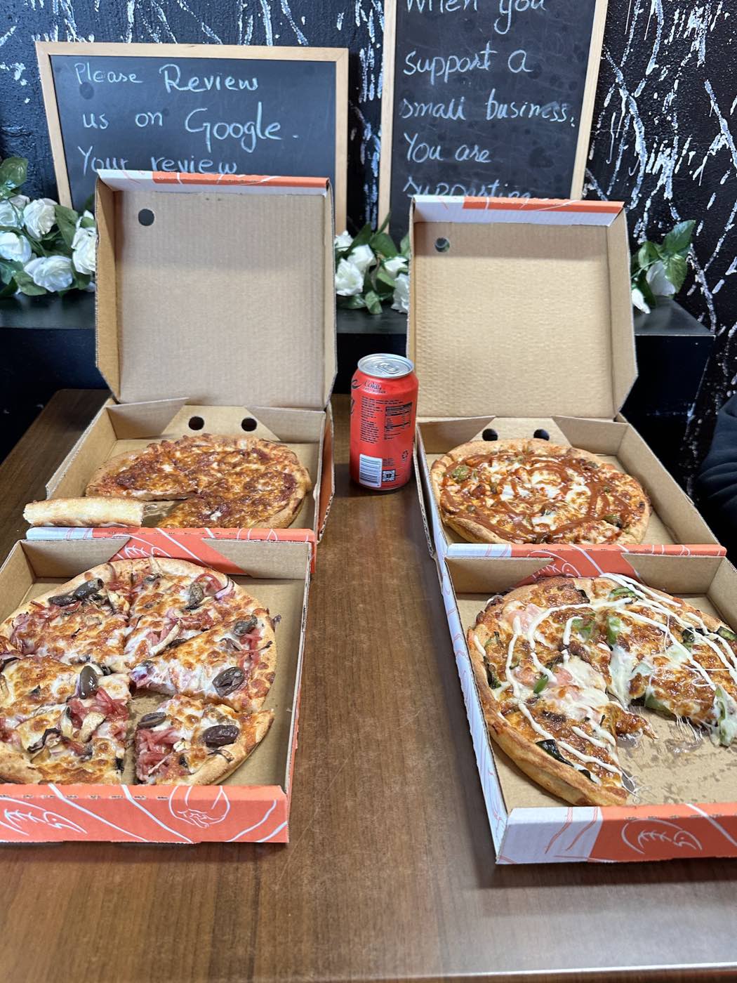 Four pizzas in open orange boxes on a table with a can of drink, in front of a chalkboard wall