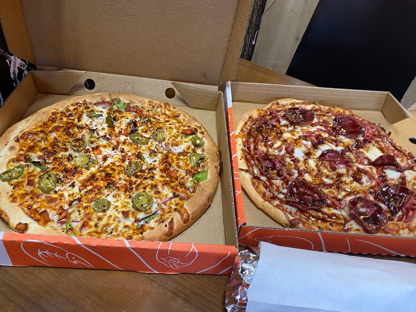Two pizzas in open orange takeaway boxes, one topped with sliced jalapenos and one with dark red meat and sauce