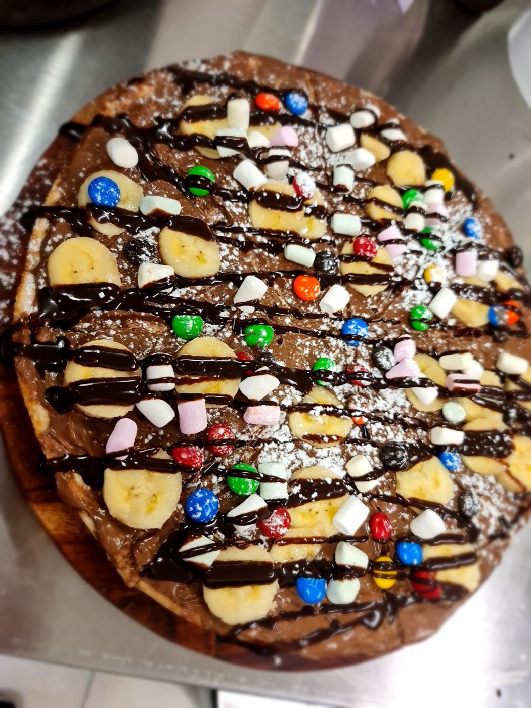 A round sweet pizza covered in banana slices, coloured chocolate sweets, marshmallows and chocolate drizzle