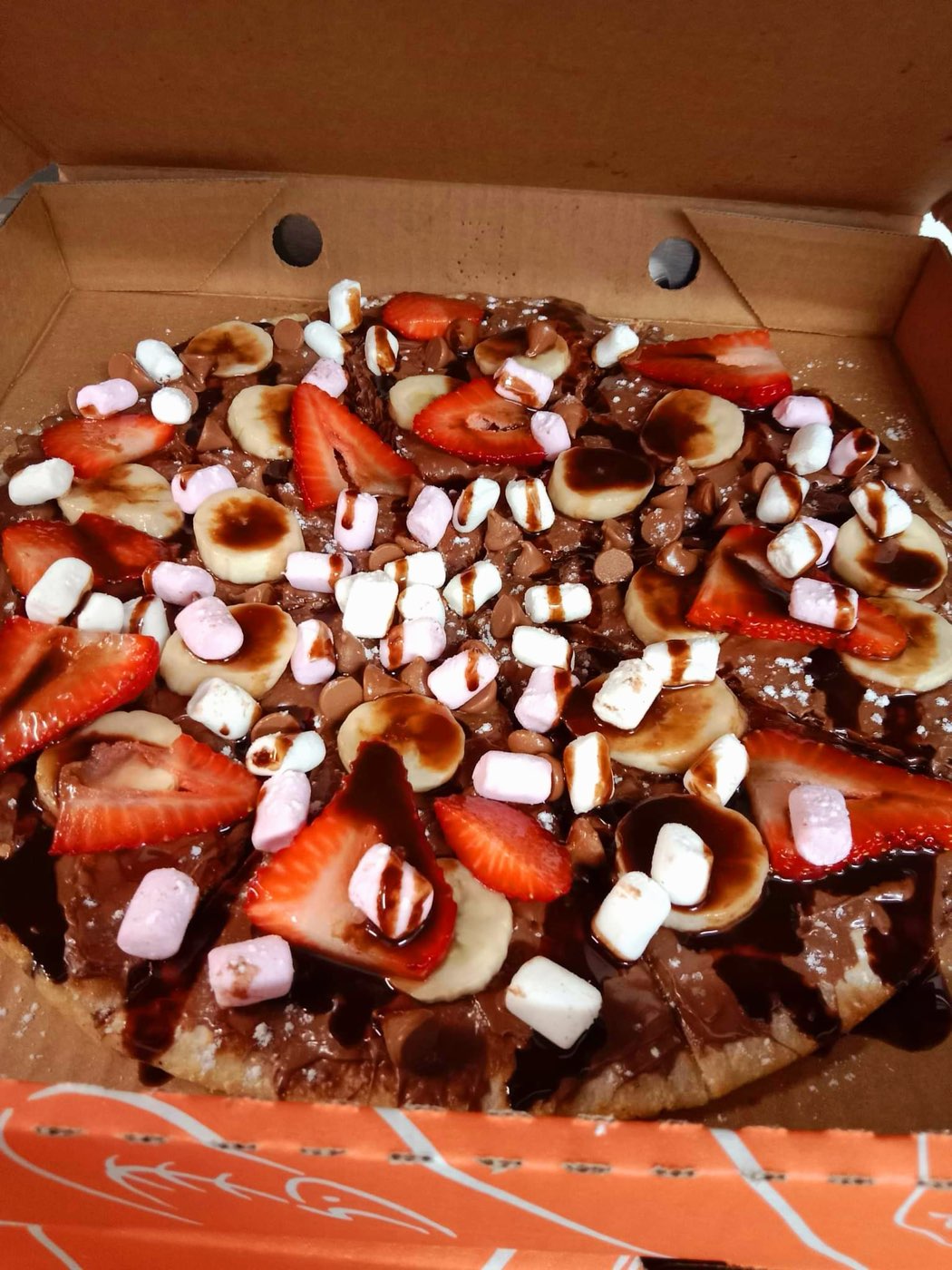 A sweet pizza in a box topped with sliced strawberries, banana, marshmallows and chocolate